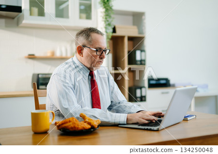 Mature Businessman using laptop while sitting at home. man sitting in kitchen Mature Businessman using laptop while sitting at home. man sitting in kitchen 134250048