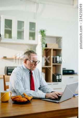 Mature Businessman using laptop while sitting at home. man sitting in kitchen and doing market research working Mature Businessman using laptop while sitting at home. man sitting in kitchen and doing market research working 134250049