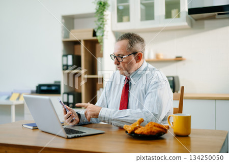 Mature Businessman using laptop while sitting at home. man sitting in kitchen and doing market research working Mature Businessman using laptop while sitting at home. man sitting in kitchen and doing market research working 134250050