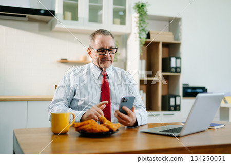 Mature Businessman using laptop while sitting at home. man sitting in kitchen Mature Businessman using laptop while sitting at home. man sitting in kitchen 134250051