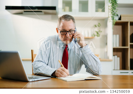 Mature Businessman using laptop while sitting at home. man sitting in kitchen Mature Businessman using laptop while sitting at home. man sitting in kitchen 134250059