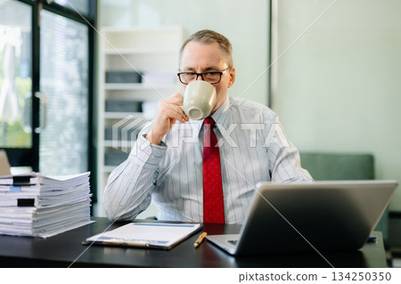 Mature businessman working in office with laptop, tablet, and notes. Perfect for business, finance, accounting 134250350