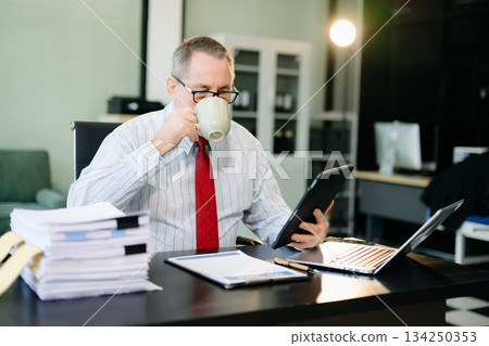 Mature businessman working in office with laptop, tablet, and notes. Perfect for business, finance, accounting Mature businessman working in office with laptop, tablet, and notes. Perfect for business, finance, accounting 134250353