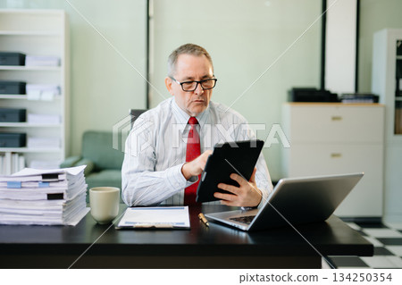 Mature businessman working in office with laptop, tablet, and notes. Perfect for business, finance, accounting 134250354