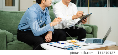 Happy Asian business partners collaborating on startup ideas using tablet and laptop in modern office. 134250487