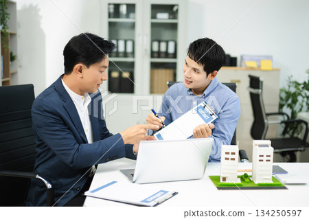 Two businessmen discuss and sign a real estate contract with housing models on a desk for concepts of property, investment Two businessmen discuss and sign a real estate contract with housing models on a desk for concepts of property, investment 134250597