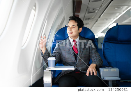 Confident businessman in first class looking out the airplane window, capturing luxury travel, success, and the modern lifestyle of business professionals. 134250981