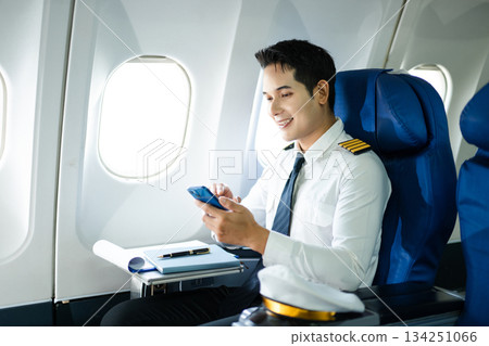 Confident pilot in uniform using smartphone in airplane cabin, ideal for aviation, communication, and travel lifestyle stock imagery. 134251066