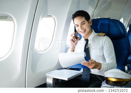 Asian Confident male pilot in uniform leaning at the passenger seat while standing inside of the airplane flight cockpit during takeoff and checking flight plan 134251072