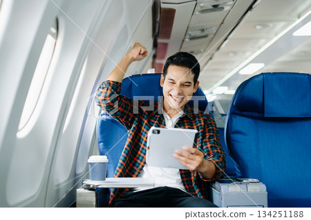 Happy young traveler celebrating victory with a fist pump on the airplane. Business success, achievement, and excited travel journey. Win, style, phone, tech. Happy young traveler celebrating victory with a fist pump on the airplane. Business success, achievement, and excited travel journey. Win, style, phone, tech. 134251188
