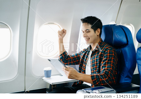 Happy young traveler celebrating victory with a fist pump on the airplane. Business success, achievement, and excited travel journey. Win, style, phone, tech. Happy young traveler celebrating victory with a fist pump on the airplane. Business success, achievement, and excited travel journey. Win, style, phone, tech. 134251189