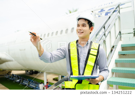 Ground crew engineer in safety vest and helmet pointing and holding documents during airplane inspection, perfect for aviation and flight safety themes Ground crew engineer in safety vest and helmet pointing and holding documents during airplane inspection, perfect for aviation and flight safety themes 134251222