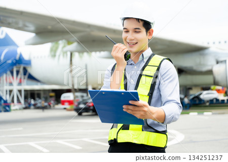 Ground crew engineer in safety vest and hard hat using walkie talkie while checking documents, perfect for aviation, maintenance, 134251237