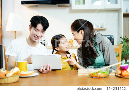 Asian family enjoys time together in kitchen as daughter uses tablet and mother prepares fresh food. Concept of parenting, home learning, and healthy lifestyle. 134251322