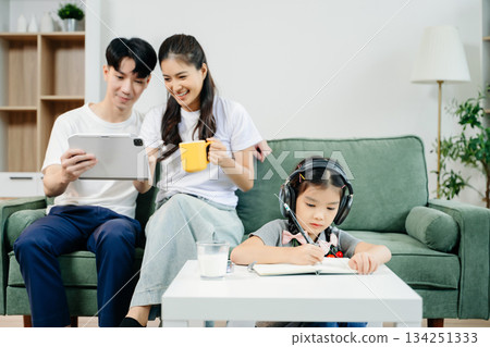 Asian child wearing headphones, studying and drawing at home, while parents enjoy coffee and tablet nearby. Family time, learning Asian child wearing headphones, studying and drawing at home, while parents enjoy coffee and tablet nearby. Family time, learning 134251333