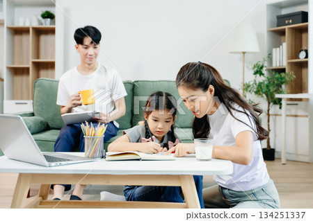 Asian parents cheering and guiding daughter through homework at home. Joyful teamwork, love, and learning in a cozy, 134251337