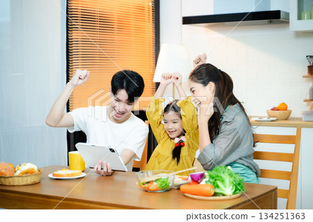 Happy Asian family cheers while using a tablet in the kitchen. Concept of digital learning, online success, family bonding, parenting, and joyful home moments. 134251363