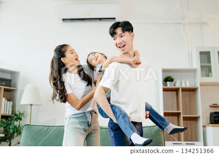Asian parents and daughter enjoying a playful piggyback ride, laughing and bonding in cozy living room. Love, happiness, and family time Asian parents and daughter enjoying a playful piggyback ride, laughing and bonding in cozy living room. Love, happiness, and family time 134251366
