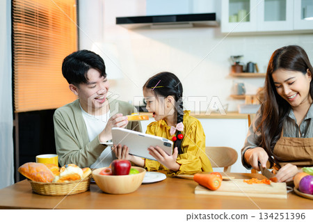 Asian family enjoys time together in kitchen as daughter uses tablet and mother prepares fresh food. Concept of parenting, home learning, and healthy lifestyle. Asian family enjoys time together in kitchen as daughter uses tablet and mother prepares fresh food. Concept of parenting, home learning, and healthy lifestyle. 134251396