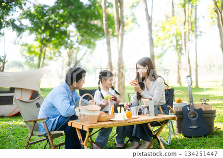 Joyful Asian family camping in a sunny garden, bonding over BBQ, tech, and laughter. Parents and daughter enjoy outdoor 134251447