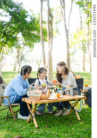 Joyful Asian family camping in a sunny garden, bonding over BBQ, tech, and laughter. Parents and daughter enjoy outdoor 134251448