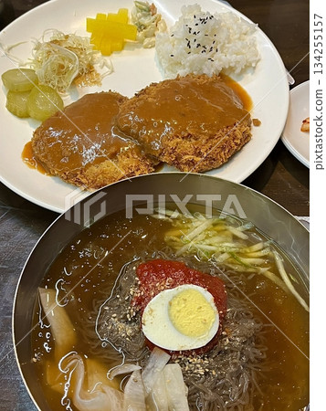 Cold naengmyeon noodles served in a chilled broth with sliced vegetables and egg, paired with crispy golden donkatsu, creating a balance of refreshing and savory flavors in one meal 134255157