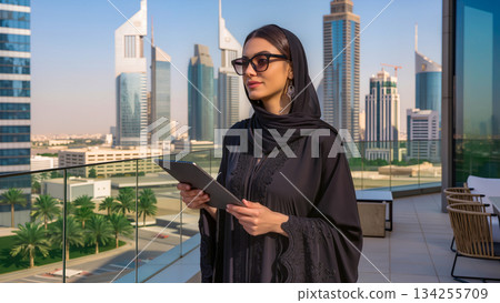 Modern Arab woman in black abaya with tablet on terrace with Dubai skyscrapers in the background. 134255709