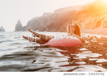 Woman, floating, sea. Young woman relaxes on a pink inflatable ring in the sea with rocky coastline. Woman, floating, sea. Young woman relaxes on a pink inflatable ring in the sea with rocky coastline. 134255742