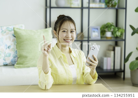 一位面帶微笑的日本女性手持智慧型手機，在客廳裡擺出「第一名」的姿勢，看向鏡頭。 134255951