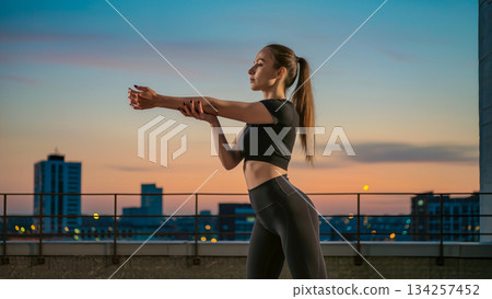 Young woman in black sportswear stretches shoulders on rooftop at sunset. Young woman in black sportswear stretches shoulders on rooftop at sunset. 134257452