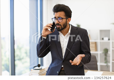 An Indian businessman wearing glasses looks concerned while talking on his phone in his office 134257624