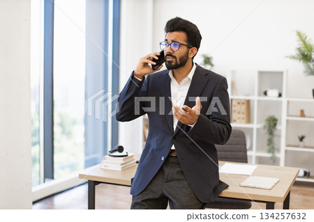 An Arabian businessman in a suit is talking on the phone in his office, looking concerned An Arabian businessman in a suit is talking on the phone in his office, looking concerned 134257632
