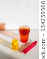 Orange glass of water, yellow vitamin bottle and pink sachet on white table 134259360