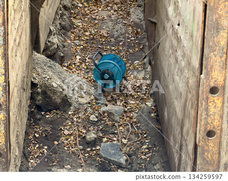Deep excavation trench featuring robust concrete shoring walls and a discarded blue industrial hose reel, symbolizing ongoing urban utility infrastructure work captured by a mobile phone 134259597