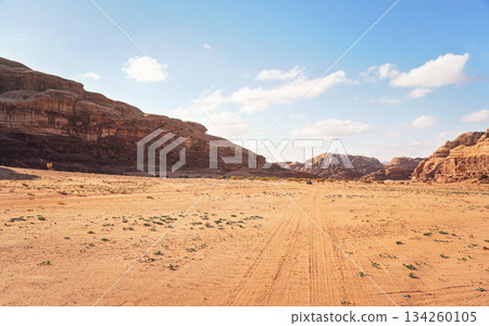 Rocky massifs on red sand desert, bright cloudy sky in background, small 4wd vehicle at distance - typical scenery in Wadi Rum, Jordan 134260105