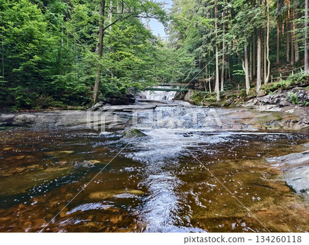 quiet river moving past rocks toward bridge, peaceful stream flowing over stones toward wooden 134260118