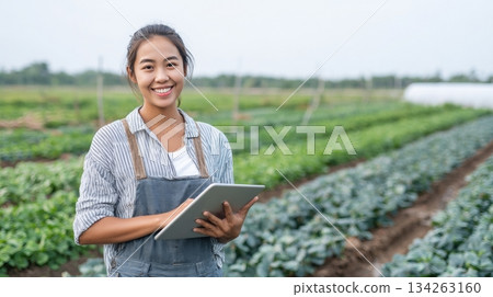 Young Southeast Asian American female farmer smiling with digital tablet in hand, surrounded by lush green vegetable rows, celebrating modern agriculture 134263160