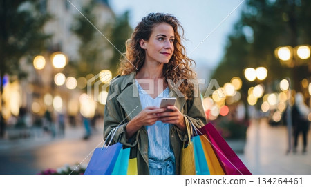 Woman shopping outdoors at sunset, holding colorful bags and checking smartphone, surrounded by bokeh street lights, creating a vibrant urban atmosphere 134264461
