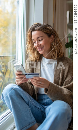 Woman sitting by bright window, happily reviewing online purchase on smartphone, cozy home ambiance with copy space for text 134264462