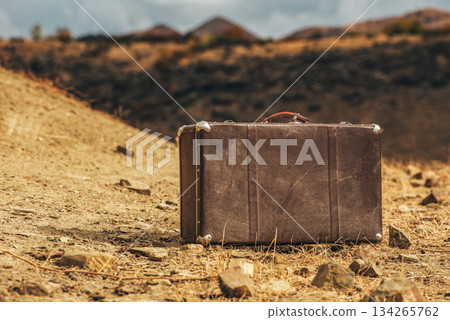 A brown suitcase is sitting on a rocky, dry ground A brown suitcase is sitting on a rocky, dry ground 134265762