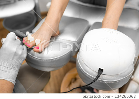 pedicure master applies gel polish to a woman toenails during procedure in a beauty salon. UV lamp for drying nail polish and gel pedicure master applies gel polish to a woman toenails during procedure in a beauty salon. UV lamp for drying nail polish and gel 134266368
