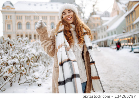 Young happy woman in a winter coat and scarf walks along a snowy city street. Concept of style. Young happy woman in a winter coat and scarf walks along a snowy city street. Concept of style. 134266787