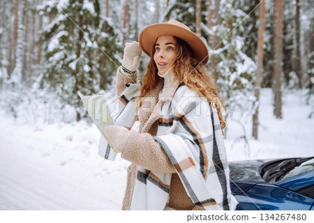 Happy woman in hat with map near the car on a snowy winter road in the forest. Car travel concept. Happy woman in hat with map near the car on a snowy winter road in the forest. Car travel concept. 134267480