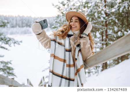 Happy woman in a hat and scarf with a phone in her hands taking a selfie in a snowy forest. 134267489