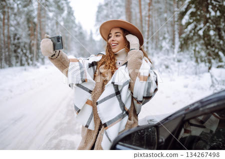 Young tourist takes a selfie with her phone next to her car on a snowy road. Travel concept. Young tourist takes a selfie with her phone next to her car on a snowy road. Travel concept. 134267498