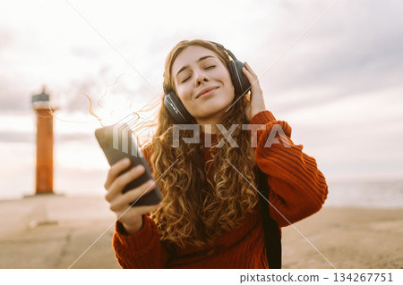 Happy woman listening nice music by waterfront while wearing headphones. Travel, blogging concept. 134267751