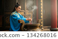 A man in traditional attire places joss sticks in a bronze container. The incense sends thin trails of smoke into the air inside a temple, creating a sacred atmosphere. 134268016