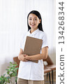 A woman in a white coat smiling and holding a binder by the window 134268344