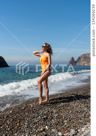 Woman swimsuit beach stands relaxing on sunny pebble shoreline near cliffs Woman swimsuit beach stands relaxing on sunny pebble shoreline near cliffs 134269239