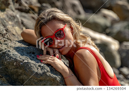 Woman, beach, vacation. A blonde woman in a red swimsuit relaxes on a sunny rocky beach. 134269304